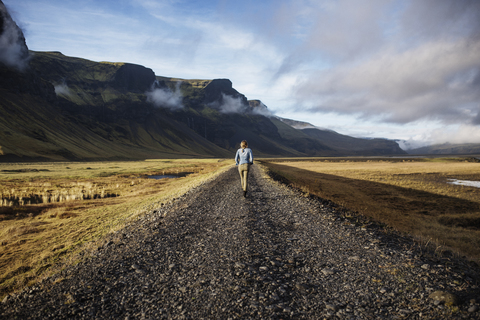 Rear view of woman walking on dirt road by mountains against sky stock photo