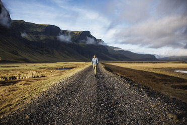 Rear view of woman walking on dirt road by mountains against sky - CAVF44621