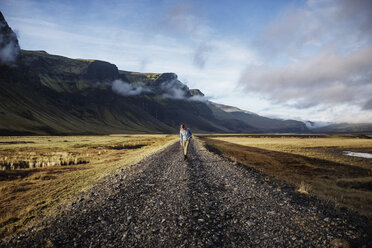 Woman walking on dirt road amidst field against mountains and sky - CAVF44622