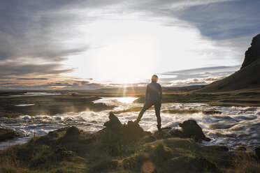 Rear view of woman standing on rocks by river against sky on sunny day - CAVF44626