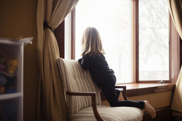 Rückansicht eines Mädchens, das zu Hause auf einem Sessel am Fenster kniet - CAVF44650