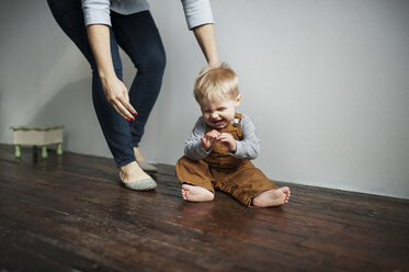 Tiefschnitt einer Mutter, die neben einem fröhlichen kleinen Jungen steht, der zu Hause auf dem Boden sitzt - CAVF44660