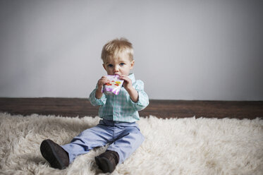 Porträt eines niedlichen kleinen Jungen, der Saft aus einer Verpackung trinkt, während er zu Hause auf einem Teppich sitzt - CAVF44662