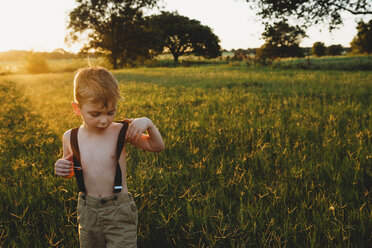 Hemdloser Junge mit Hosenträgern, der bei Sonnenuntergang inmitten von Pflanzen auf einem Feld steht - CAVF44690