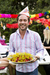 Porträt eines glücklichen erwachsenen Mannes, der einen Teller mit gekochtem Hummer hält, mit Freunden im Hintergrund - MASF06058