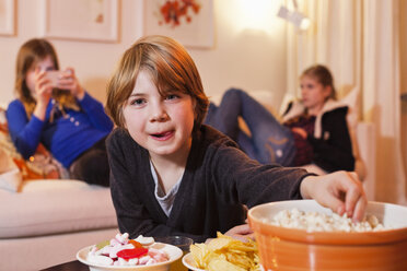 Porträt eines kleinen Jungen, der am Kaffeetisch Popcorn isst, mit Schwestern im Hintergrund - MASF06116