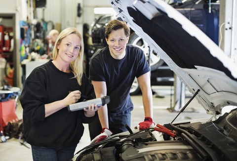 Porträt eines glücklichen erwachsenen weiblichen Mechanikers, der ein digitales Tablet hält und mit einem Kollegen an einer offenen Motorhaube steht, lizenzfreies Stockfoto