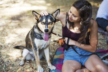 Smiling woman stroking dog while sitting at park - CAVF44705