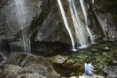 Rückansicht einer Frau, die auf einem Felsen vor einem Wasserfall sitzt - CAVF44774