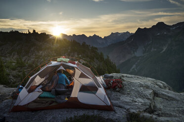 Frau sitzt im Zelt auf einer Klippe gegen Berge bei Sonnenuntergang - CAVF44776