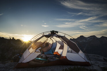 Rückansicht einer im Zelt sitzenden Frau gegen die Berge bei Sonnenuntergang - CAVF44777