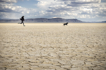 Frau und Hund laufen auf einem trockenen Feld vor bewölktem Himmel in der Wüste von Alvord - CAVF44780