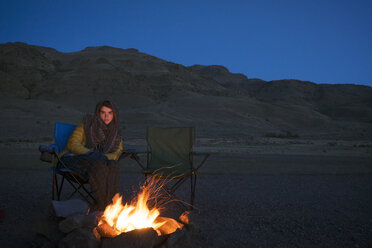Porträt einer Frau, die auf einem Stuhl am Lagerfeuer sitzt, mit Blick auf Berge und blauen Himmel in der Wüste von Alvord - CAVF44783