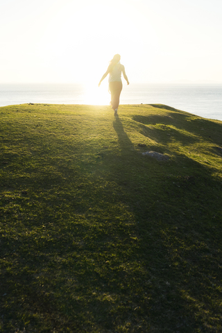 Rückansicht einer Frau, die bei Sonnenuntergang auf einem grasbewachsenen Hügel am Meer spazieren geht, lizenzfreies Stockfoto