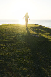 Rückansicht einer Frau, die bei Sonnenuntergang auf einem grasbewachsenen Hügel am Meer spazieren geht - CAVF44792