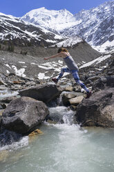 Seitenansicht von Frau springt auf Felsen am Fluss gegen schneebedeckte Berge - CAVF44796