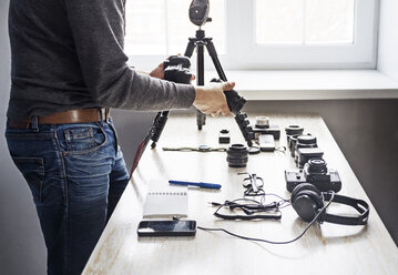 Midsection of male photographer holding lens and camera by table - CAVF44830