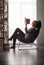 Frau hält ein Buch in der Hand und entspannt sich auf einem Stuhl am Fenster zu Hause - CAVF44838