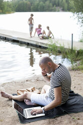 Älterer Mann, der einen Laptop benutzt, während er am Strand mit seiner Familie auf dem Pier sitzt und mit dem Handy kommuniziert - MASF06222