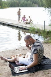 Mature man using laptop while communicating on cell phone at beach with family sitting on pier - MASF06222