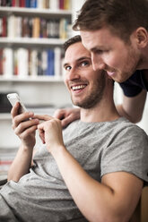 Glücklicher junger schwuler Mann, der seinen Partner ansieht, während er zu Hause ein Mobiltelefon benutzt - MASF06331