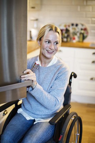 Glückliche behinderte Frau mit Flasche beim Öffnen der Kühlschranktür in der Küche, lizenzfreies Stockfoto