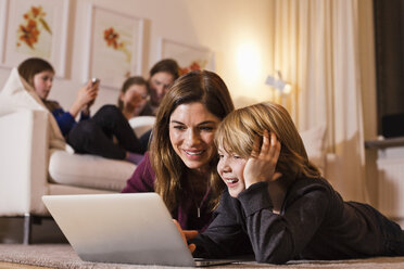 Glückliche Mutter und Sohn benutzen einen Laptop auf dem Boden, während die Familie auf dem Sofa im Hintergrund sitzt - MASF06383