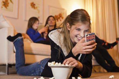 Glückliches Mädchen, das ein Mobiltelefon benutzt, während es Popcorn auf dem Boden isst, während die Familie auf dem Sofa im Wohnzimmer sitzt, lizenzfreies Stockfoto