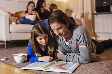 Father and daughter reading map on floor with family sitting on sofa in living room - MASF06454