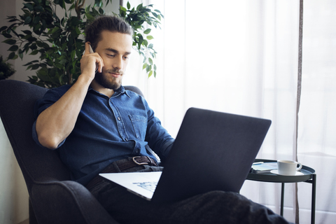 Mann spricht mit einem Mobiltelefon, während er zu Hause einen Laptop benutzt, lizenzfreies Stockfoto