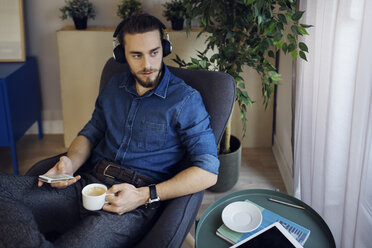 Mann hält Kaffeetasse und Mobiltelefon und hört Musik, während er zu Hause auf einem Stuhl sitzt - CAVF45018