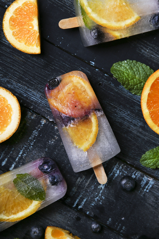 Hausgemachtes Detox-Eis mit Heidelbeeren, Orangenscheiben und Minzblättern auf schwarzem Holz, lizenzfreies Stockfoto