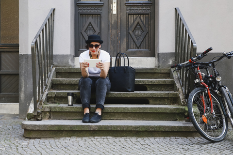 Frau sitzt auf der Treppe mit Kaffee zum Mitnehmen und benutzt ein Tablet, lizenzfreies Stockfoto