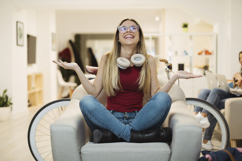 Happy casual young woman sitting in armchair in coworking space - OCAF00218