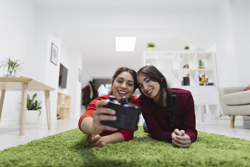 Zwei glückliche junge Frauen, die in einem Coworking Space auf dem Boden liegen und ein Selfie machen - OCAF00245