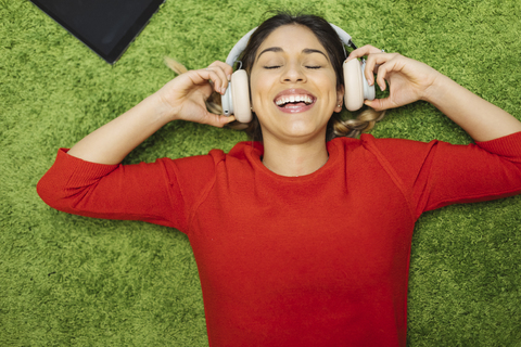 Glückliche junge Frau, die auf einem Teppich liegt und mit Kopfhörern Musik hört, lizenzfreies Stockfoto