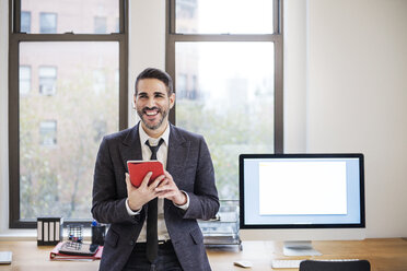 Happy businessman using tablet computer while leaning on desk in office - CAVF45337