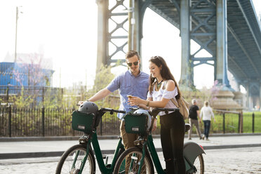 Glückliches Paar schaut auf das Telefon, während es mit Fahrrädern vor der Manhattan-Brücke steht - CAVF45355