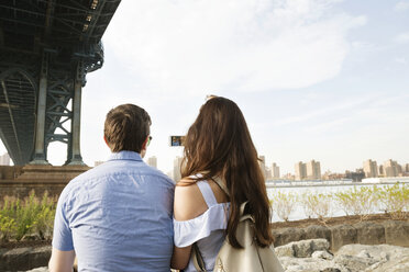 Rückansicht eines Paares, das ein Telefon benutzt, während es unter der Manhattan-Brücke gegen den Himmel sitzt - CAVF45361