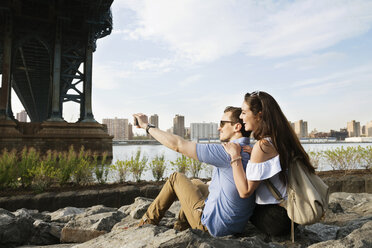 Glückliches Paar, das ein Selfie macht, während es auf den Felsen am East River gegen den Himmel sitzt - CAVF45362