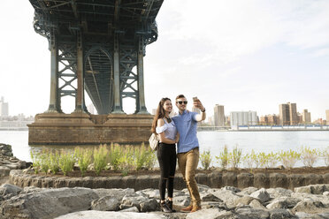 Paar, das ein Selfie macht, während es auf den Felsen unter der Manhattan Bridge gegen den klaren Himmel steht - CAVF45363
