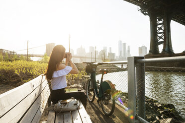 Frau beim Fotografieren auf einer Bank an der Manhattan-Brücke gegen den klaren Himmel sitzend - CAVF45365