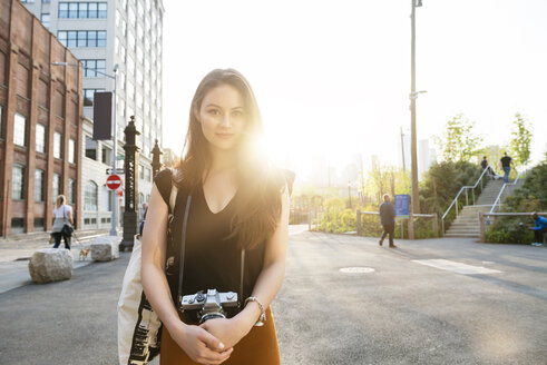 Porträt einer jungen Frau mit Kamera, die auf einer Straße in der Stadt vor einem klaren Himmel steht - CAVF45369