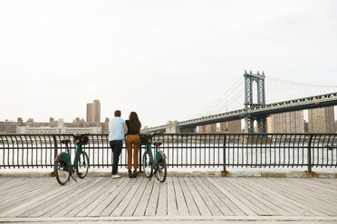 Couple standing with bicycles on footpath by Manhattan bridge against sky - CAVF45371