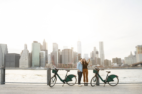 Paar, das ein Selfie macht, während es neben Fahrrädern am East River in der Stadt steht, lizenzfreies Stockfoto