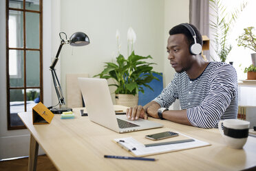 Junger Mann hört Musik, während er im Büro zu Hause einen Laptop benutzt - CAVF45530