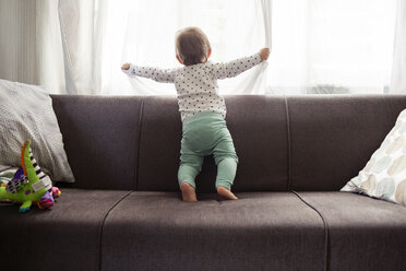 Ein kleines Mädchen schaut durch das Fenster, während es auf dem Sofa zu Hause steht - CAVF45562