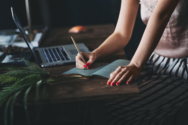 Midsection of woman writing in book by laptop on table - CAVF45667