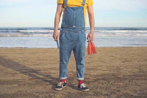 Mann in Latzhose steht am Strand und hält Flaschen mit Erfrischungsgetränken, lizenzfreies Stockfoto