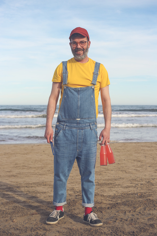 Porträt eines lächelnden Mannes in Latzhose, der am Strand steht und Flaschen mit Erfrischungsgetränken hält, lizenzfreies Stockfoto